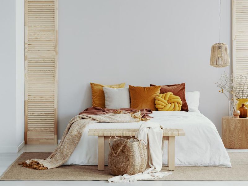 Brown and orange pillows on white bed in natural bedroom interior with wicker lamp and wooden bedside table with vase