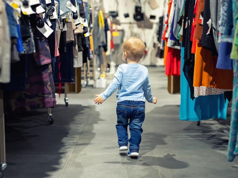 Cute caucasian blond toddler boy walking alone at clothes retail store between rack with hangers. Baby discovers adult shopping world. Baby get lost at big hypermarket shopping mall