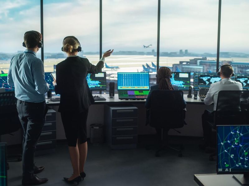 Female and male air traffic controllers with headsets talk in airport tower while computers display airplane departure and arrival data on navigation screens