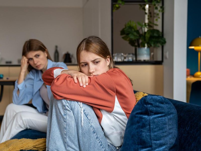 Frustrated offended teenage girl sits hugging knees and looks away. Mom sits next daugther