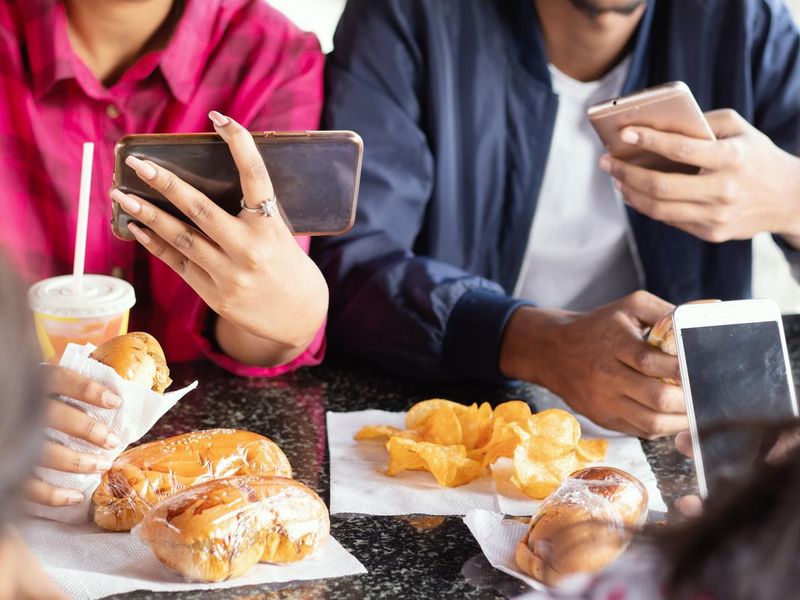 Group of unrecognizable people busy on mobile while having snacks together at restaurant - Concept of millennials having junk food, phone addiction, social media sharing and connection.
