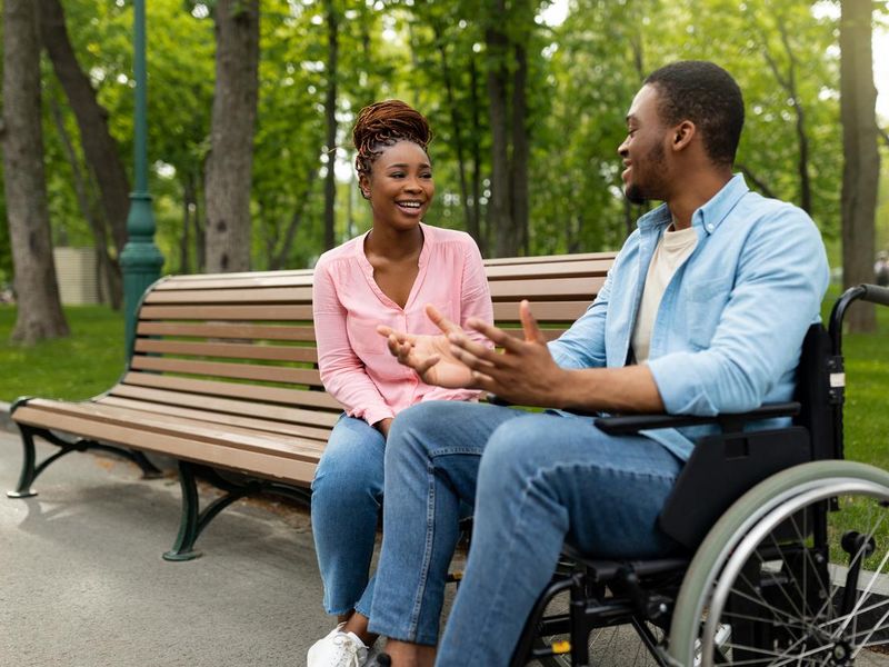 Happy handicapped black guy in wheelchair talking to his girlfriend at park, copy space