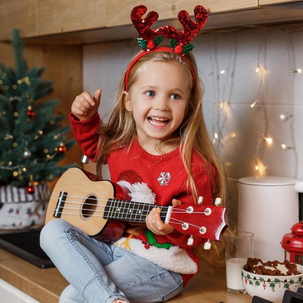 Happy little girl in red horns playing ukulele on the background of christmas tree. Talent kid having fun on winter holidays at cozy decorated home kitchen.