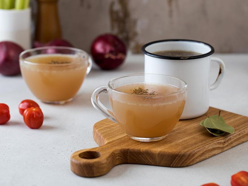Homemade bone broth in mugs with spices on a wooden stick with bay leaf, tomatoes and onions.