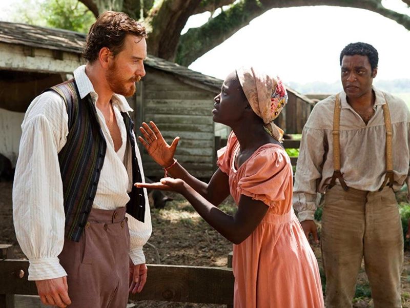 Lupita Nyong'o speaking to Michael Fassbender with Chiwetel Ejiofor looking on in 12 Years a Slave