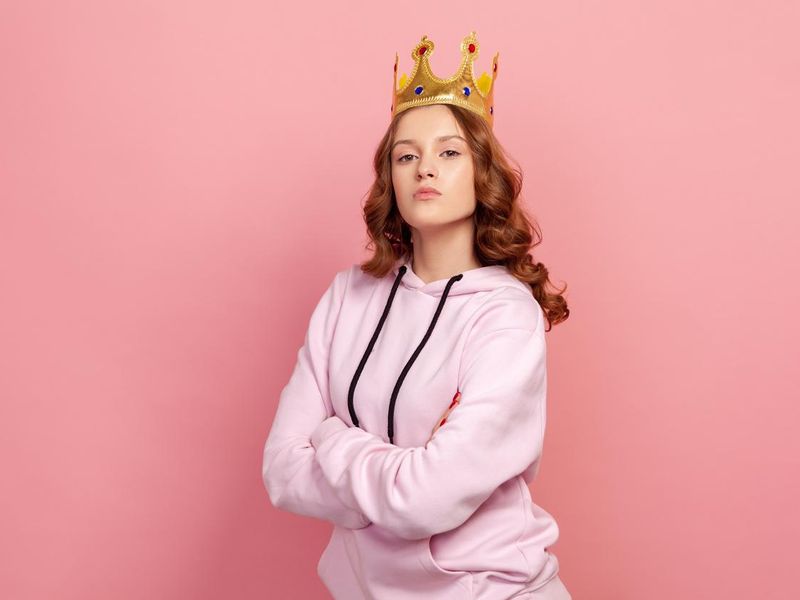 Portrait of confident curly haired teenage girl in hoodie with gold crown on head crossing hands and looking at camera