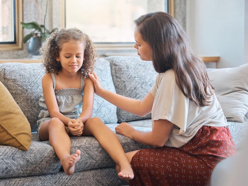 Sad little girl sitting on couch while mother tries to talk to her