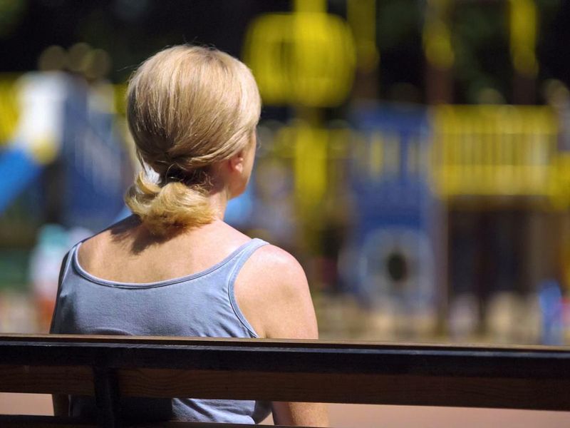 Sad woman sitting on playground bench and looking at children