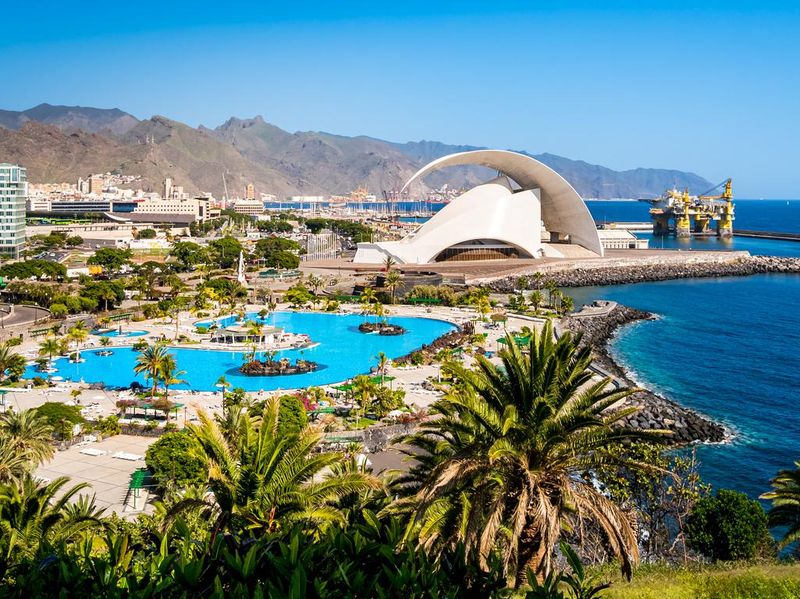 Santa Cruz de Tenerife with iconic Auditorio de Tenerife concert hall shining in the sunlight against a backdrop of Anaga mountains, an oil rig and a tropical pool with palm trees in the foreground.