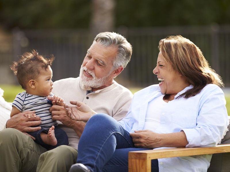 Senior couple sitting in the garden with their baby grandson, smiling at him, front view