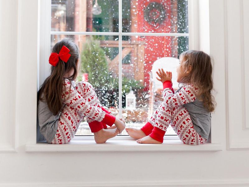 Two cute girls in pajamas sitting and looking out the window at snowy weather. Christmas moments with kids at home