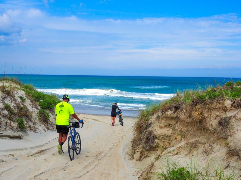 Two people pushing their bike down a sandy beach access with a deserted shore and waves crashing