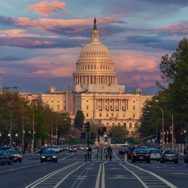 West Façade of the US Capitol Building as seen from Pennsylvania Avenue, Washington DC, USA. Spectacular Red, Orange and Blue Sunset Sky is in background. Cars, Bicyclists, Traffic lights, Green Trees and American flag are in the image.