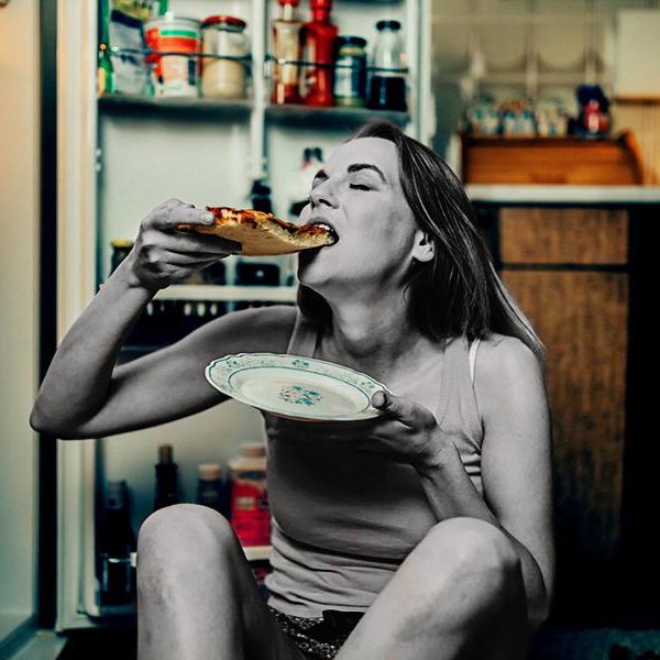 Young casual dressed woman eating pica slice in front of the refrigerator late night in the kitchen