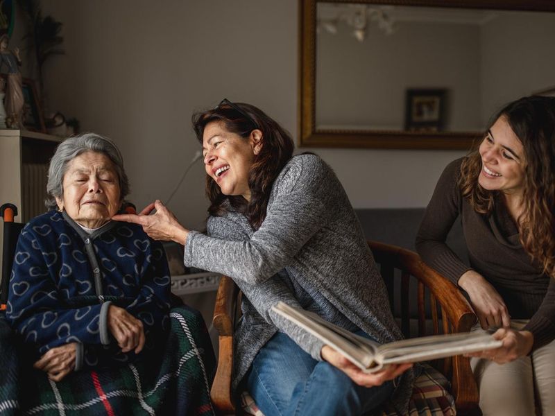 women generation with old sick grandmother sitting in wheelchair and smiling daughter and granddaughter looking a photo album.