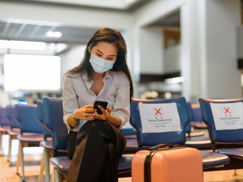 Young Asian tourist woman with mask using phone and sitting with distance at the airport
