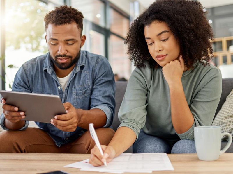 Young mixed race couple going through documents and using a digital tablet at a table together at home. Hispanic husband and wife planning and paying bills. Boyfriend and girlfriend filling in forms
